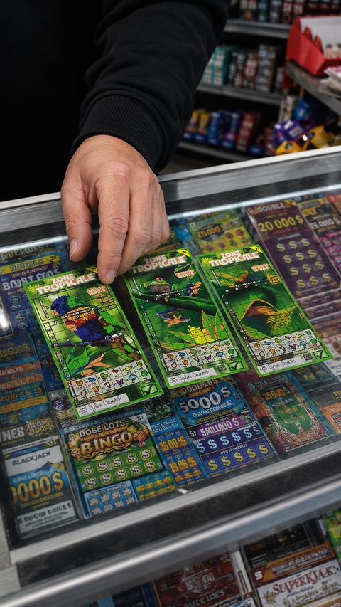 Une main tient un billet à gratter Zone Tropicale de Loto-Québec au-dessus d’un présentoir de billets.